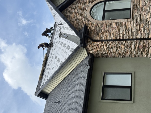 Two roofers on a steep gable laying underlayment on a stone-fronted house