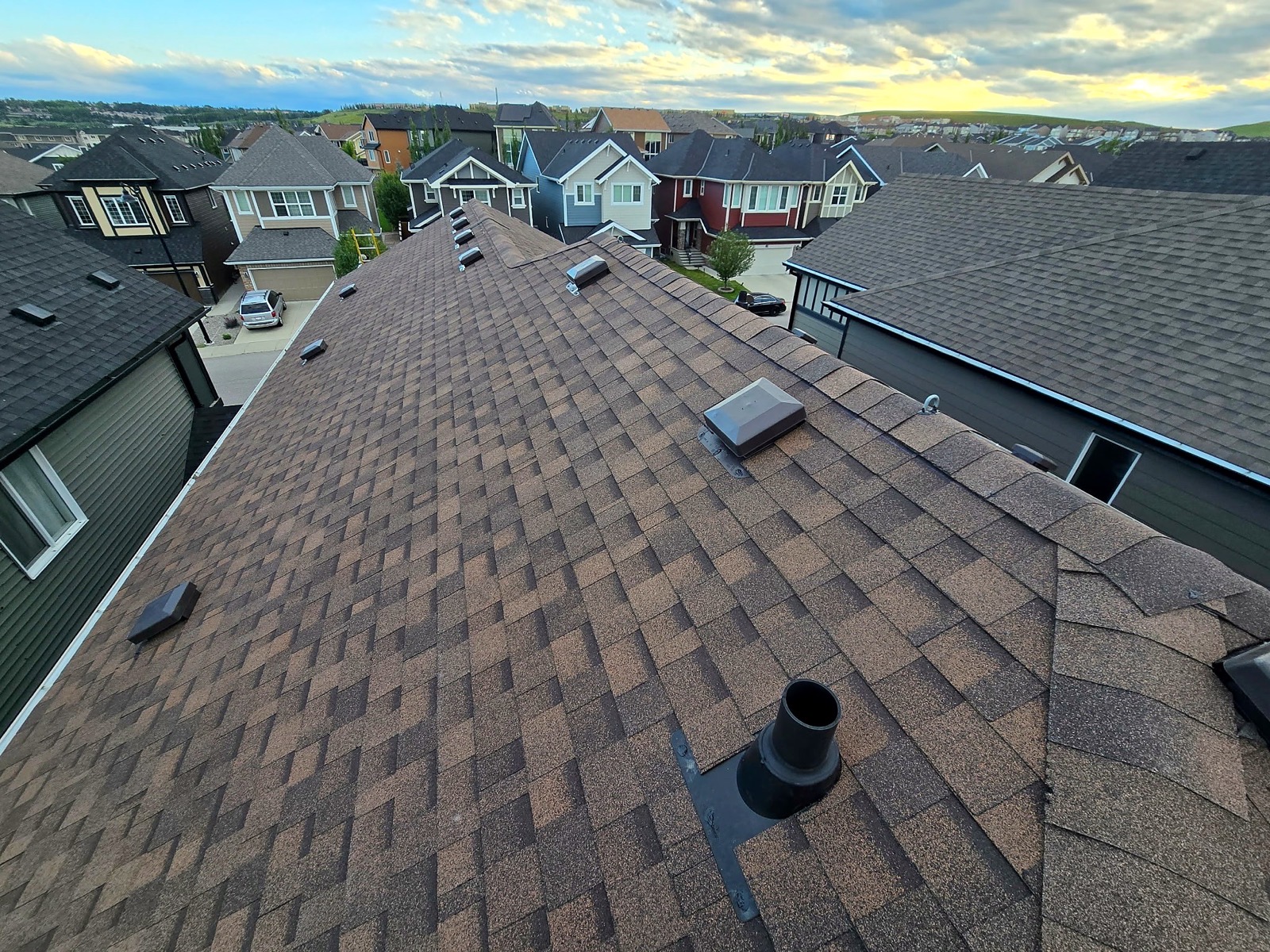 Brown asphalt shingle roof with vents and pipe flashing, houses along the street beyond, evening sky